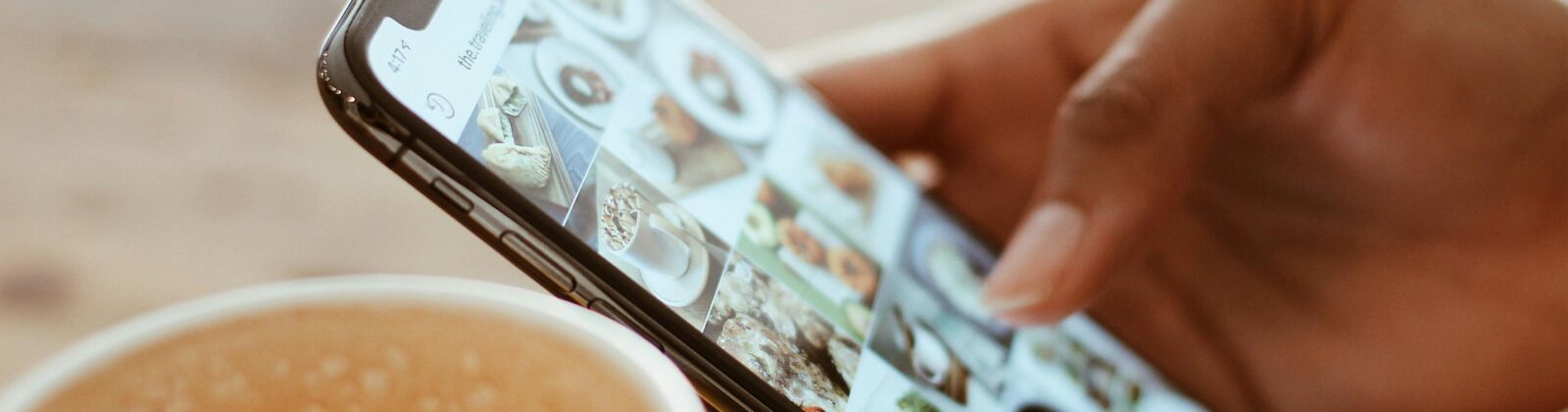 Hand browsing social media photos on a smartphone next to a cup of coffee indoors.