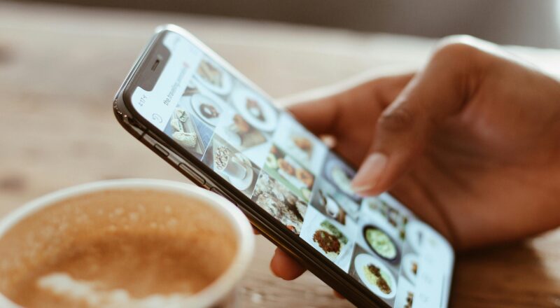 Hand browsing social media photos on a smartphone next to a cup of coffee indoors.