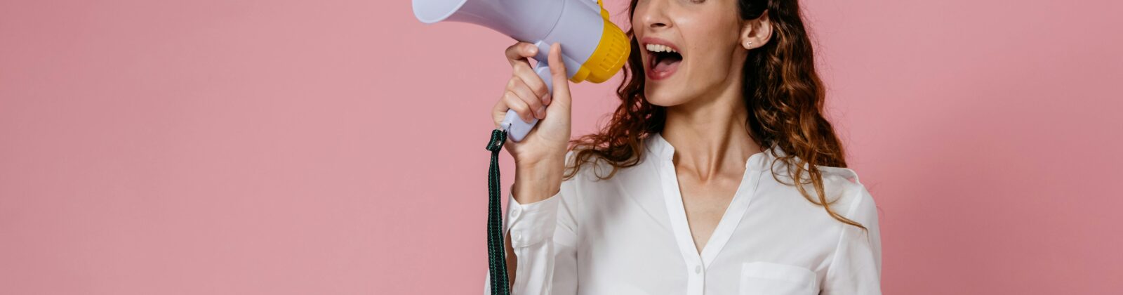 Woman in white shirt holding megaphone against a pink background, confidently speaking.